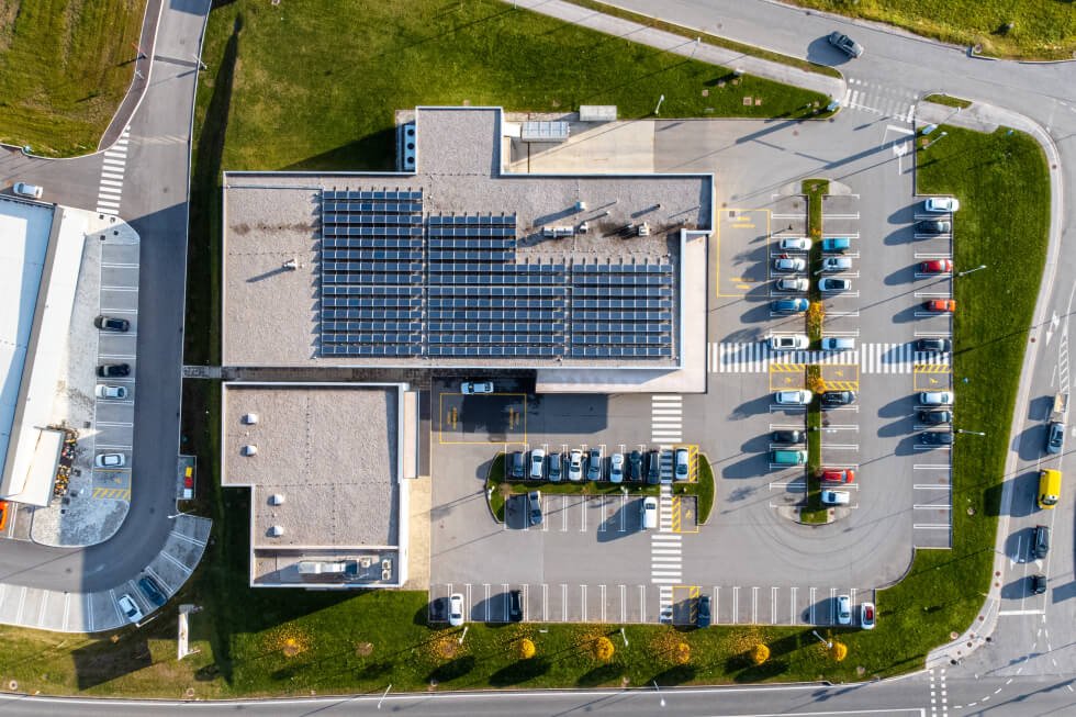 Aerial view of a commercial building with solar panels installed on top through a commercial tax strategy purchase. The building has a partially full parking lot and is surrounded by green grass.